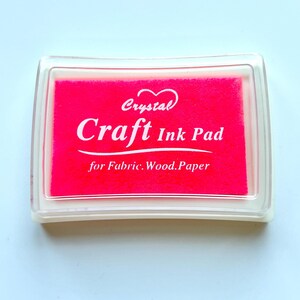 K&ouml;nnte beinhalten: Ein rosa Stempelkissen mit dem Text "Crystal Craft Ink Pad for Fabric, Wood, Paper" in wei&szlig;.