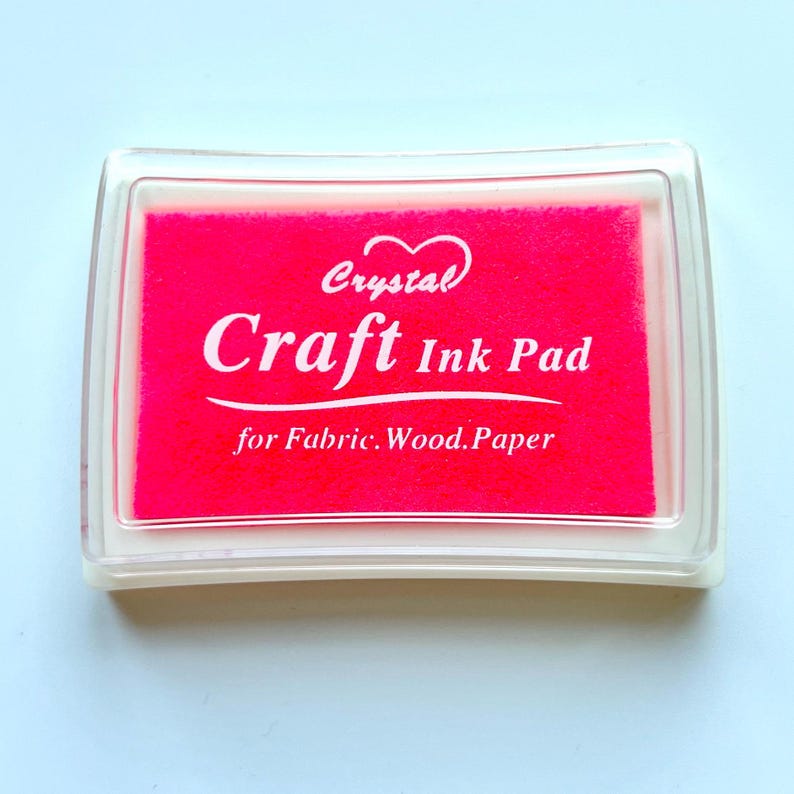 K&ouml;nnte beinhalten: Ein rosa Stempelkissen mit dem Text "Crystal Craft Ink Pad for Fabric, Wood, Paper" in wei&szlig;.