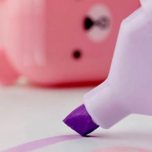 May include: A purple highlighter with a white barrel is drawing a line on a white surface. The highlighter is in focus, while a pink highlighter with a cartoon bear face is out of focus in the background.
