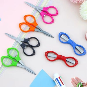 Può includere: Un assortimento di forbici e taglierine colorate. Le forbici hanno lame in acciaio inossidabile e manici rosa, arancioni, neri e verdi. Le taglierine sono blu e rosse. Sono visibili anche una penna multicolore e un piccolo portachiavi con forbici verdi.
