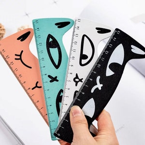 May include: Three colorful rulers with cat-face designs. The rulers are peach, teal, and black, each with a different expression. The rulers measure up to 15 inches and are held by a hand.