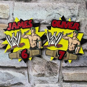 May include: Two WWE themed cake toppers featuring a wrestler with a championship belt. The toppers are yellow and black with the wrestler's name, "JAMES" and "OLIVER", in red. The numbers "6" and "7" are also included on the toppers.