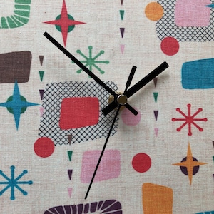 Puede incluir: Una esfera de reloj blanca con agujas negras y un centro dorado. La esfera del reloj tiene un diseño retro con formas geométricas coloridas y estrellas.