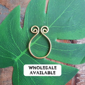 May include: Gold wire ring with a unique design, featuring two loops at the top and a teardrop shape. The ring is displayed on a green leaf. The text "Wholesale Available" is printed on a white label.