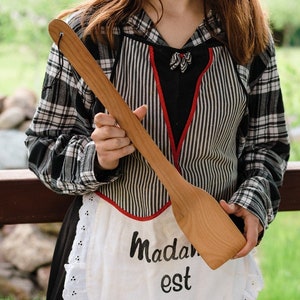 May include: A long, light-colored wooden spatula with a rounded end and a hole in the handle. The spatula is held in front of a person wearing a black and white plaid shirt and a white apron with the text "Madam est".
