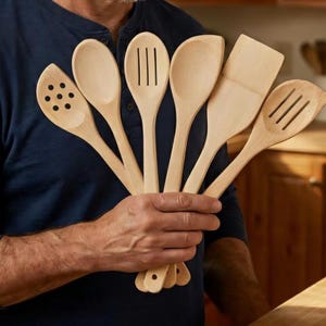 May include: A set of six light-colored wooden kitchen utensils arranged in a fan shape. The collection includes a perforated spoon, a solid spoon, a slotted spoon, a curved spoon, a flat spatula, and a slotted turner, all with smooth handles.