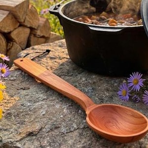 May include: A long-handled wooden spoon with a smooth, reddish-brown grain finish and a small hole at the end of the handle. The spoon is positioned near a black cast iron pot containing a stew and several small purple and yellow wildflowers.