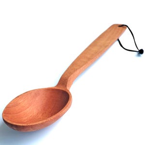 May include: A wooden spoon with a long handle and a rounded bowl. The spoon is made of light brown wood and has a smooth finish. A black string is attached to the end of the handle for hanging. Ideal for cooking and serving.