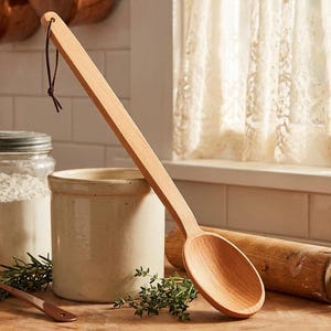 May include: A long-handled wooden cooking spoon with a smooth, light-toned finish and a dark leather hanging loop attached to the end of the handle. The spoon features a deep, oval-shaped bowl and a straight, flat handle for a comfortable grip.