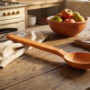 May include: A long-handled wooden ladle with a smooth, light brown finish and a rounded bowl. The handle features a small hole at the end with a thin, dark brown leather cord threaded through it, resting on a light-colored fabric cloth.