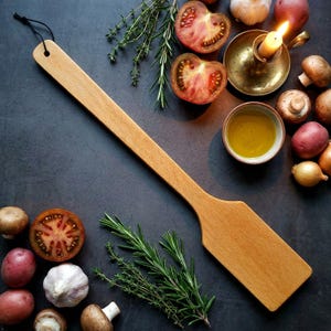 May include: A long, flat wooden spatula with a smooth, light brown finish and a small circular hole at the end of the handle for hanging. The utensil features a wide, rectangular blade and a slender, elongated handle, crafted from solid wood.