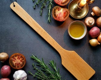 May include: A long, flat wooden spatula with a smooth, light brown finish and a small circular hole at the end of the handle for hanging. The utensil features a wide, rectangular blade and a slender, elongated handle, crafted from solid wood.