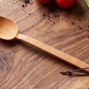 May include: A light brown wooden spoon with a smooth, oval-shaped bowl and a long, flat handle. A thin, dark brown leather cord is threaded through a small hole at the end of the handle, allowing for easy hanging storage of the kitchen utensil.