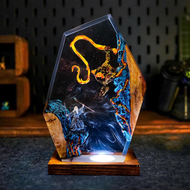May include: A unique resin art piece featuring a fantasy scene with a wizard, a fiery creature, and blue and orange accents. The artwork is encased in a clear, geometric shape and set on a wooden base, illuminated by a soft light.