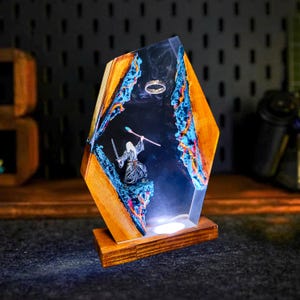 May include: A unique wooden and resin lamp featuring a fantasy scene. The lamp has a geometric shape with wooden sides and a clear resin center. Inside, a miniature figure of a wizard stands with a sword and staff, with a ring floating above. The lamp is illuminated from within.