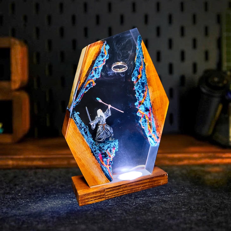 May include: A unique wooden and resin lamp featuring a fantasy scene. The lamp has a geometric shape with wooden sides and a clear resin center. Inside, a miniature figure of a wizard stands with a sword and staff, with a ring floating above. The lamp is illuminated from within.