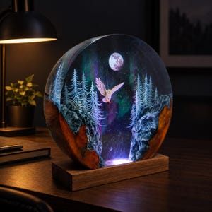 Könnte beinhalten: Eine kreisförmige Lampe aus Harz und Holz mit einer nächtlichen Waldszene, einer fliegenden weißen Eule, einem Vollmond und grünen Nordlichteffekten. Das Objekt steht auf einem rechteckigen Holzsockel und wird von unten durch eine LED beleuchtet.