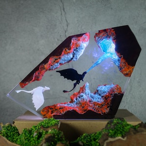 Op de afbeelding: Een decoratief kunstwerk van hars met een zwart draak silhouet en een wit draak silhouet tegen een achtergrond van vurige oranje en blauwe elementen. Het kunstwerk heeft een heldere, geometrische vorm met een lichtbron.