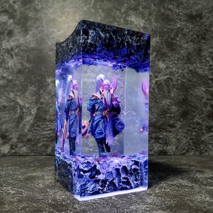 Op de afbeelding: Een rechthoekige hars sculptuur met figuren in blauwe en paarse kleding, ingekapseld in een helder blok. De sculptuur heeft een donkere, getextureerde bovenkant en een gloeiend paars interieur, wat een fantasiethema creëert.