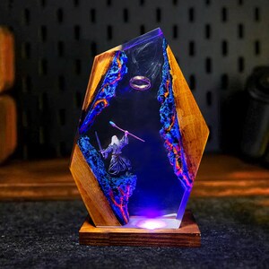 May include: A unique wooden and resin lamp featuring a fantasy scene. The lamp has a wooden frame with a clear resin center. Inside, a miniature figure of a wizard stands before a ring, with blue and orange accents.