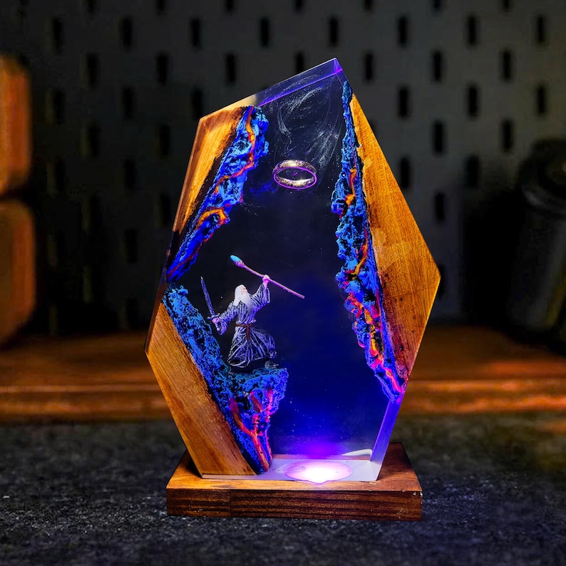 May include: A unique wooden and resin lamp featuring a fantasy scene. The lamp has a wooden frame with a clear resin center. Inside, a miniature figure of a wizard stands before a ring, with blue and orange accents.
