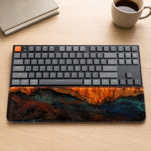 May include: A mechanical keyboard with dark gray keys and a single orange escape key, paired with a rectangular wrist rest featuring a polished wood and resin design that displays a swirling pattern of deep orange, blue, and black colors.