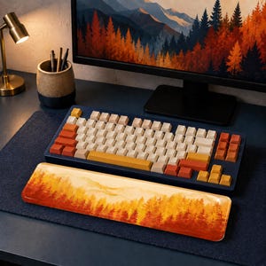 May include: A mechanical keyboard with a dark blue frame and a set of cream, orange, and yellow keycaps. In front of the keyboard sits a rectangular wrist rest featuring a printed landscape of a forest with autumn-colored trees in shades of red.