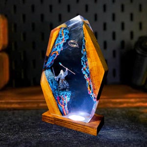 May include: A unique resin and wood lamp featuring a detailed scene with a wizard figure, a ring, and a mountain landscape. The lamp has a wooden base and a geometric shape, with blue and orange accents.