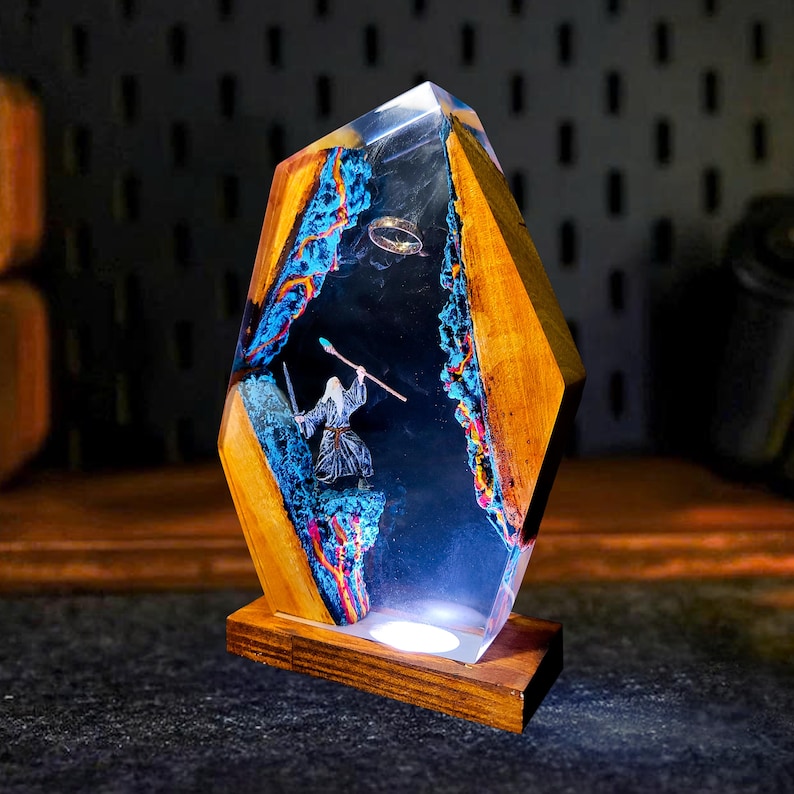 May include: A unique resin and wood lamp featuring a detailed scene with a wizard figure, a ring, and a mountain landscape. The lamp has a wooden base and a geometric shape, with blue and orange accents.