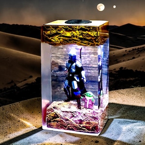 May include: A rectangular resin lamp featuring a Mandalorian figure in silver armor and a small green creature in a pink robe standing on a textured base. The top section has a rough, gold-colored finish, and the clear body illuminates the figures.