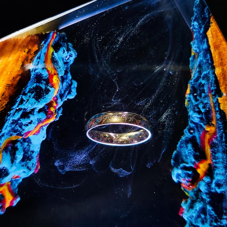 May include: A gold-colored ring with engraved text, set against a dark background with blue and orange abstract art. The ring has a glowing white outline, creating a mystical effect. The ring is a fantasy-themed item.