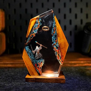 May include: A decorative resin and wood lamp featuring a scene with a wizard, a ring, and a staff. The lamp has a wooden base and a geometric shape with a clear resin center. The interior scene has blue, orange, and black accents.