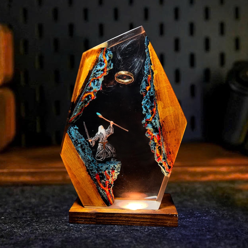 May include: A decorative resin and wood lamp featuring a scene with a wizard, a ring, and a staff. The lamp has a wooden base and a geometric shape with a clear resin center. The interior scene has blue, orange, and black accents.