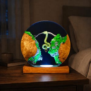 May include: A circular resin lamp featuring a central green snake hanging from a branch between two pieces of natural light-coloured wood. The scene is accented with bright green moss-like textures and a dark blue background, set on a wooden base.