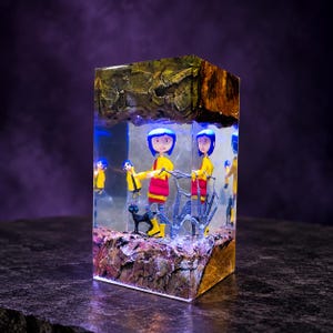 May include: A rectangular resin block featuring a miniature figure of a blue-haired character in a yellow raincoat and red boots, accompanied by a black cat. The scene is encased between textured, dark, rock-like top and bottom layers.
