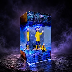 Puede incluir: Un bloque rectangular de resina con base y parte superior de madera, que encapsula una escena en miniatura con dos figuras con impermeables amarillos y pantalones azules, una de pie y otra sentada, junto a un gato negro, todo en resina azul.