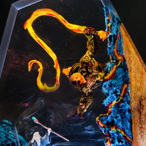 May include: A detailed fantasy diorama featuring a fiery dragon and a wizard. The dragon, with yellow and orange flames, is perched on a blue and brown rock formation. A wizard with a staff is in the lower portion of the image, set against a dark, starry background.