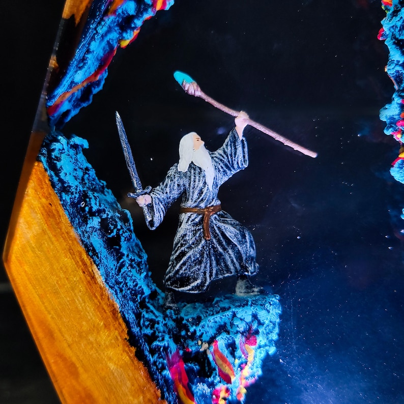 May include: A miniature wizard figure with a sword and staff, set within a resin art piece. The artwork features vibrant blue, orange, and red accents, creating a fantasy scene. The wizard is dressed in a gray robe with a brown belt.