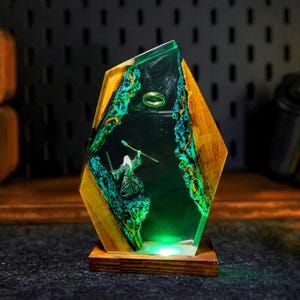 May include: A decorative resin and wood lamp featuring a miniature scene with a wizard, a ring, and a staff. The lamp has a wooden base and a geometric shape with green and blue accents. The lamp is illuminated with a green light.