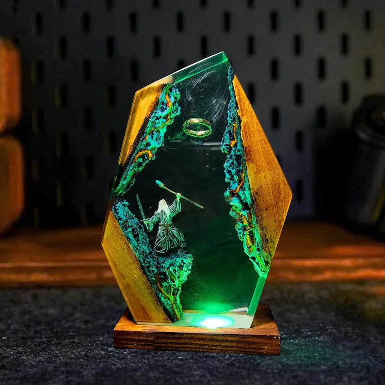 May include: A decorative resin and wood lamp featuring a miniature scene with a wizard, a ring, and a staff. The lamp has a wooden base and a geometric shape with green and blue accents. The lamp is illuminated with a green light.