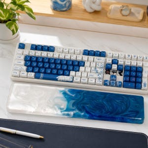 Könnte beinhalten: Eine mechanische Tastatur mit weißen und blauen Tasten mit komplizierten Blumenmustern, gepaart mit einer rechteckigen Handgelenkauflage aus Harz, die ein wirbelndes, weißes und tiefblaues Marmordesign zeigt, das an Meereswellen erinnert.