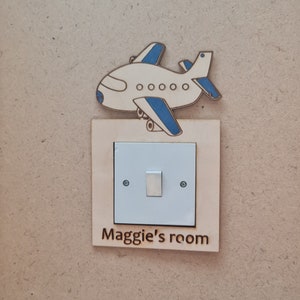 May include: A wooden light switch cover with a white light switch and a small airplane cutout above it. The cover has the text "Maggie's room" printed on it.