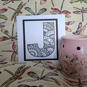 Può includere: Stampa artistica in bianco e nero della lettera "J" con motivi intricati, incorniciata in nero. A destra, un bruciatore di olio in ceramica rosa con decorazioni floreali, su tessuto a motivi di libellule.