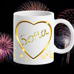 May include: White ceramic mug featuring the name "Sofia" in gold script within a gold heart outline. The mug has a white handle and is set against a black background with colorful fireworks.