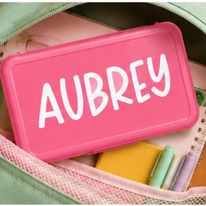 May include: A rectangular hot pink plastic pencil case with the name AUBREY printed in white capital letters on the lid, resting inside an open light green backpack that contains a spiral notebook, a yellow notepad, and three colored markers.
