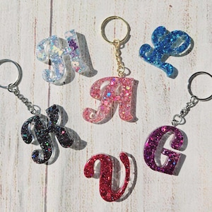 May include: Seven sparkly letter keychains with silver key rings. The letters are A, G, H, K, L, U, and V. The letters are made of clear resin with glitter embedded in the resin. The letters are in a cursive font.