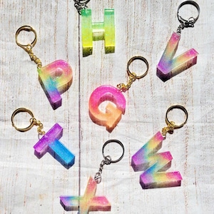 May include: Eight rainbow glitter resin letter keychains. The letters are H, P, G, T, X, V, and W. The keychains are on a white wooden background.