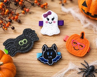 May include: Four felt hair clips featuring Halloween designs: a white ghost with a purple bow, a black spider with green eyes, a black bat with blue wings, and an orange pumpkin wearing pink glasses, each attached to a small ribbon-covered clip.