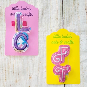 May include: Two hair clips with the letters "LC" and "FT" on pink and yellow cards. The "LC" clip is purple with a watercolor design, and the "FT" clip is pink with white stitching. The cards read "little luda's arts & crafts."