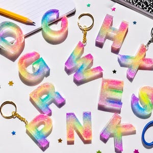 May include: A collection of resin alphabet letters featuring a rainbow gradient of pink, yellow, green, and blue colors with embedded glitter. Some letters are attached to gold or silver metal keychains, while others are loose, scattered among crayons.